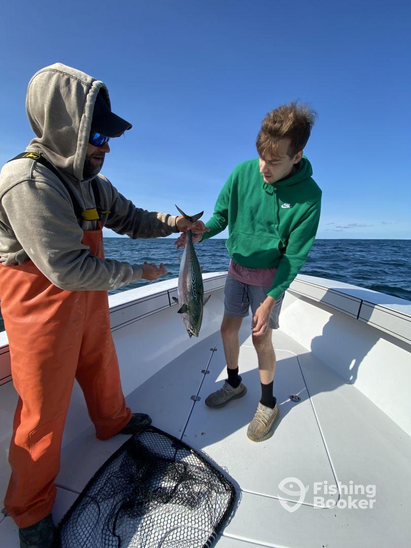A young angler and a guide on a boat, showcasing a freshly caught fish, likely a Mackerel, during a fishing trip.