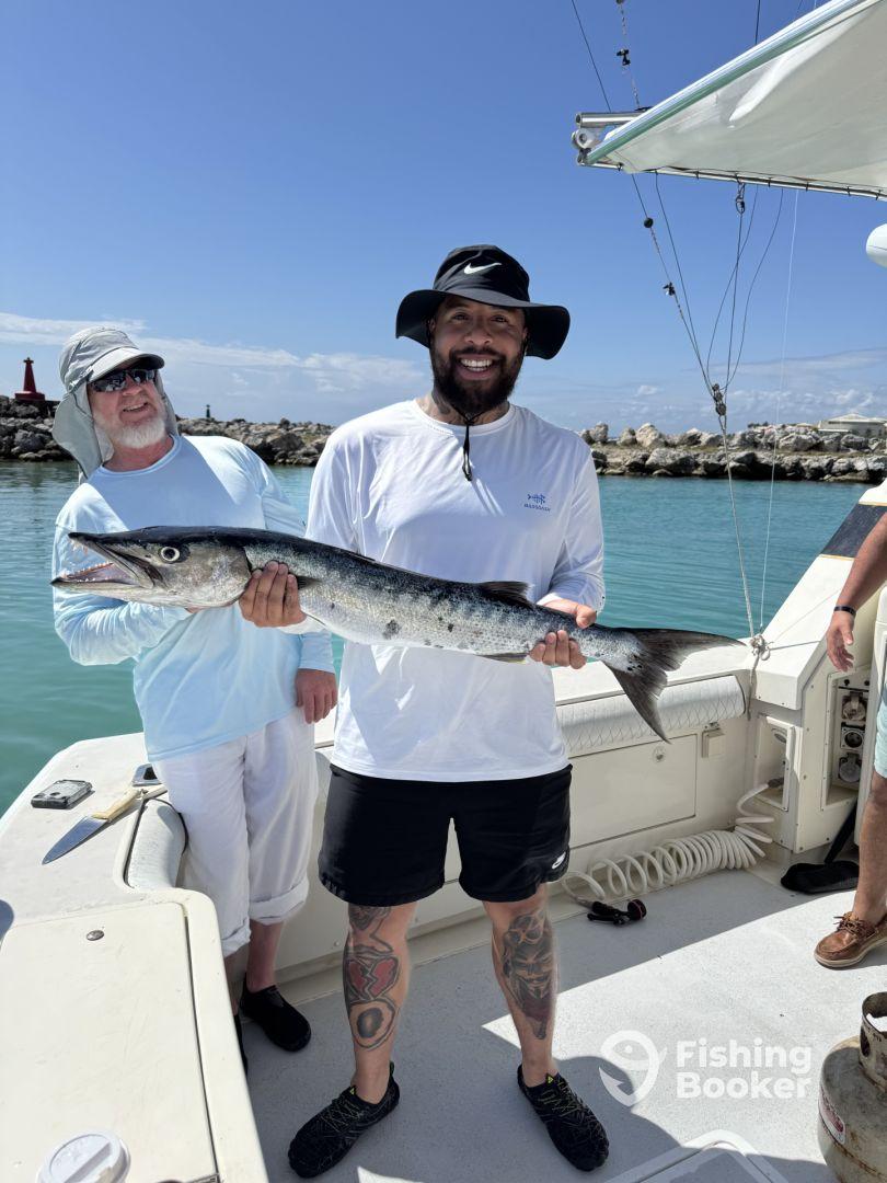 Two anglers proudly displaying a large Barracuda on a boat after a successful fishing trip.