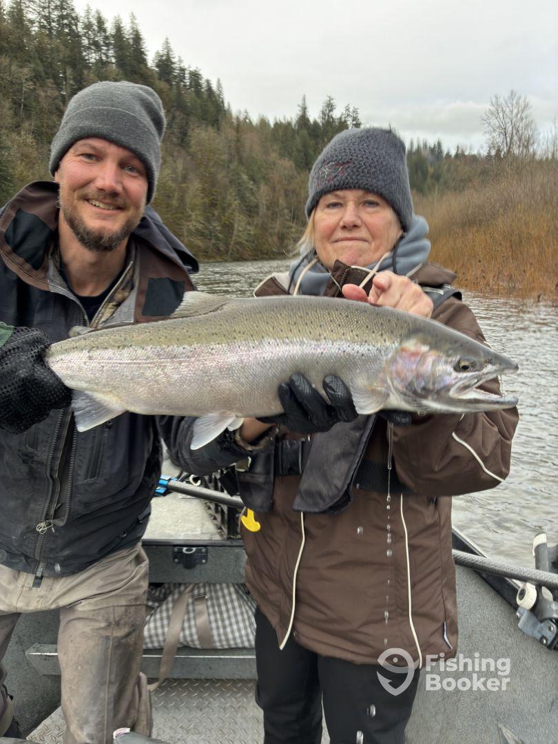 Two anglers proudly displaying a large Steelhead caught during a river fishing trip, surrounded by a scenic natural backdrop.